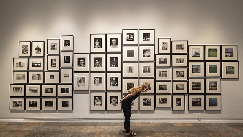 Una mujer joven vestida de negro observa detenidamente una gran instalación fotográfica compuesta por múltiples retratos en una sala de exposiciones.