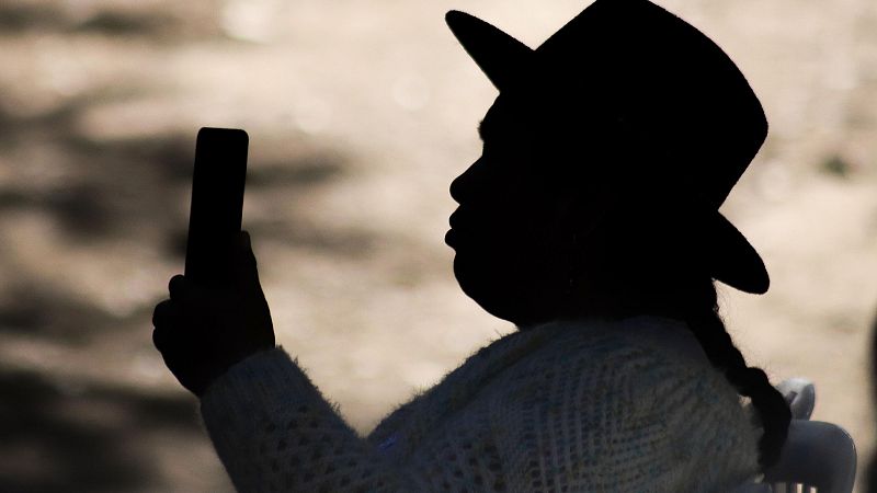 Silueta de perfil de una mujer con sombrero tradicional y trenza que sostiene un teléfono móvil frente a su rostro contra la luz del fondo.