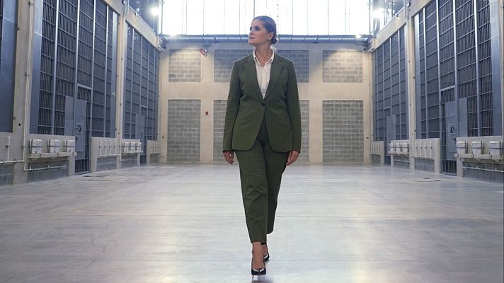 Laura Fernández camina con traje verde oliva por un pasillo entre celdas en una moderna prisión.