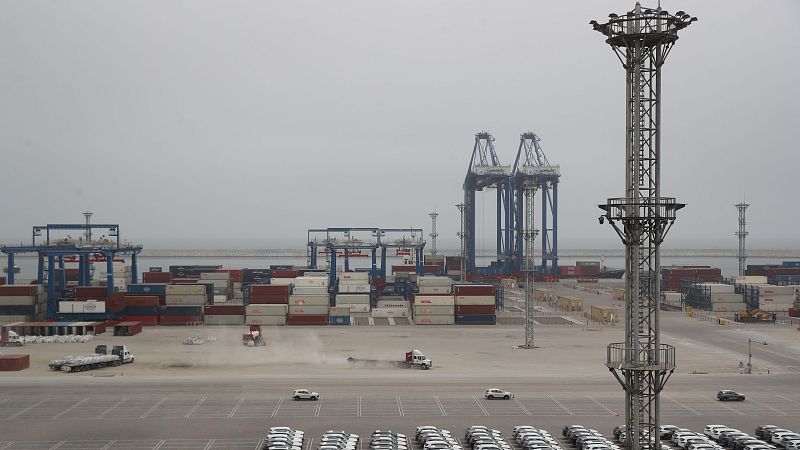 Vista panorámica del puerto de Chancay con explanada de autos nuevos, camiones, contenedores apilados, grúas pórtico y torres de iluminación bajo un cielo nublado.