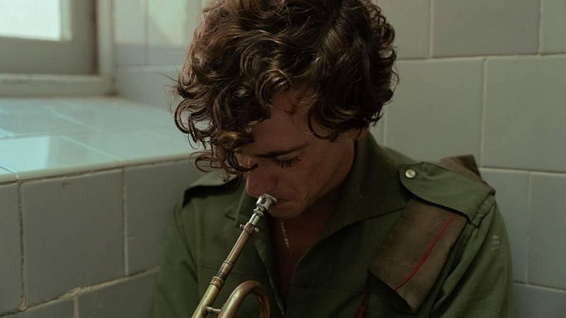 Plano medio de un joven soldado con camisa militar verde oliva y gorra en la hombrera, ensayando en silencio con una corneta de lat�n en un ba�o con azulejos.