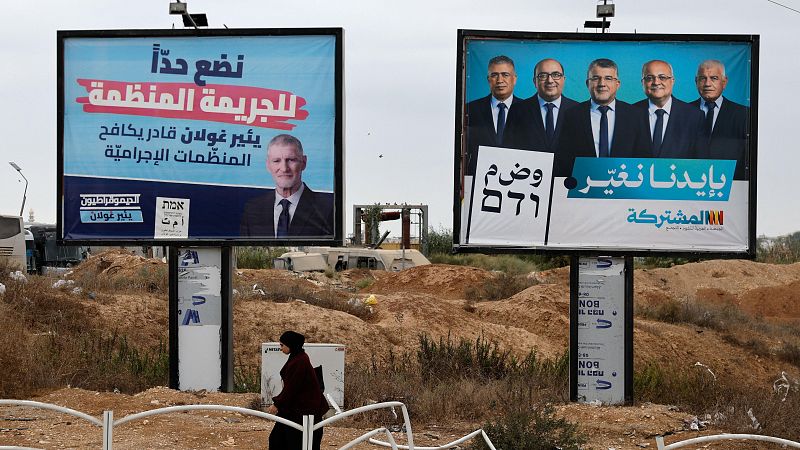 Dos grandes vallas publicitarias electorales con retratos de políticos y lemas en un terreno árido junto a una carretera, con una mujer con hiyab caminando en primer plano.