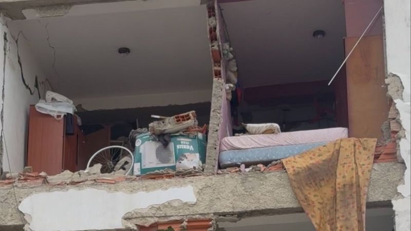 Plano frontal del derrumbe parcial de un edificio que deja al descubierto dos habitaciones con escombros, una bicicleta y una cama al borde del vacío.