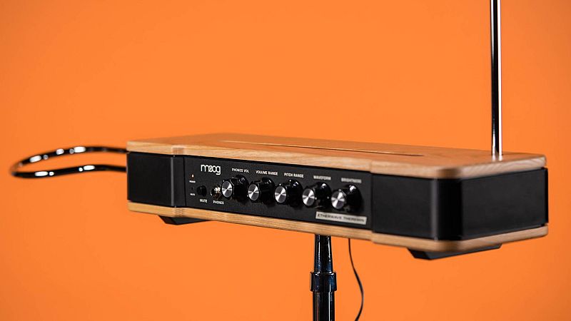 Therem�n Moog Etherwave sobre soporte con cuerpo de madera clara y chasis negro, antenas cromadas y mandos de control frontales sobre fondo naranja.