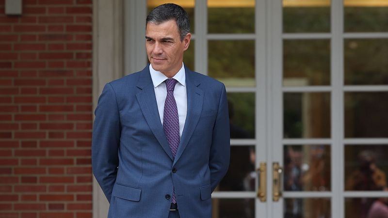 Plano medio de Pedro S�nchez en el Palacio de la Moncloa, con traje formal y expresi�n reflexiva ante la fachada del edificio.