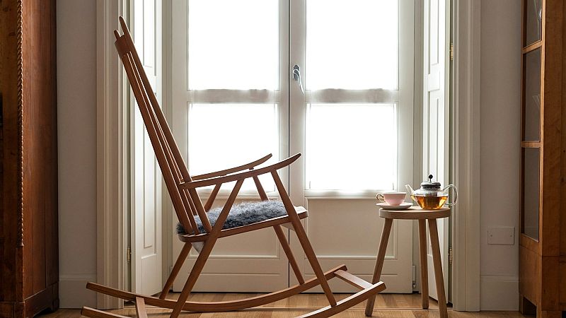 C�lido rinc�n interior con una mecedora de madera, un coj�n gris y una mesa auxiliar con tetera y taza frente a un balc�n luminoso.