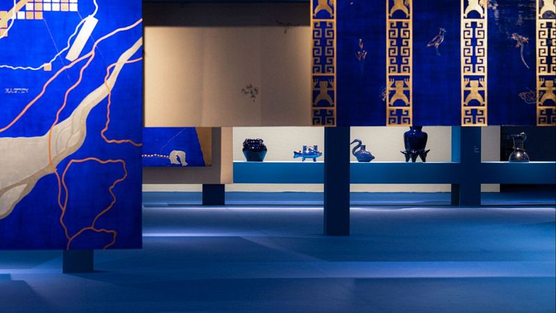 Instalación artística inmersiva en tonos azules y dorados con paneles flotantes, motivos geométricos y esculturas de cerámica vidriada sobre una bancada.