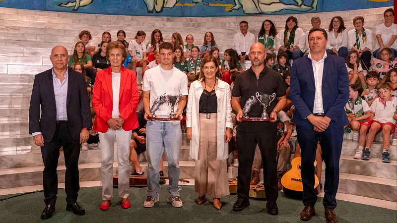 Seis personas posan en un escenario durante un acto deportivo en Cantabria, incluyendo a la presidenta regional y a representantes del Real Racing Club sosteniendo trofeos con una bota y una copa.