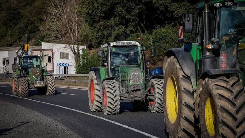 Columna de tractores agr�colas circulando por una carretera asfaltada durante una protesta del sector agrario, con un veh�culo verde de llantas rojas avanzando en primer plano.