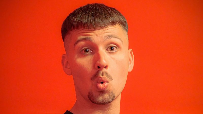 Primer plano de un joven con bigote y perilla, cabello casta�o y expresi�n de asombro sobre fondo rojo.