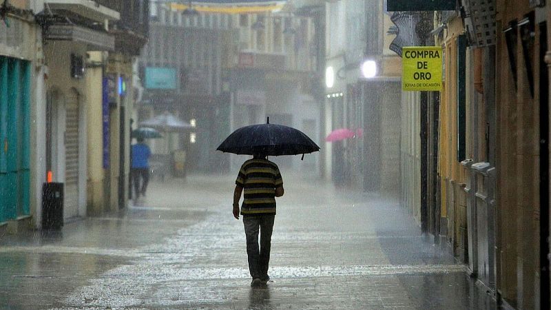 Calle peatonal urbana y h�meda bajo la lluvia intensa, con un viandante de espaldas portando un paraguas negro y un cartel de compra de oro a la derecha.