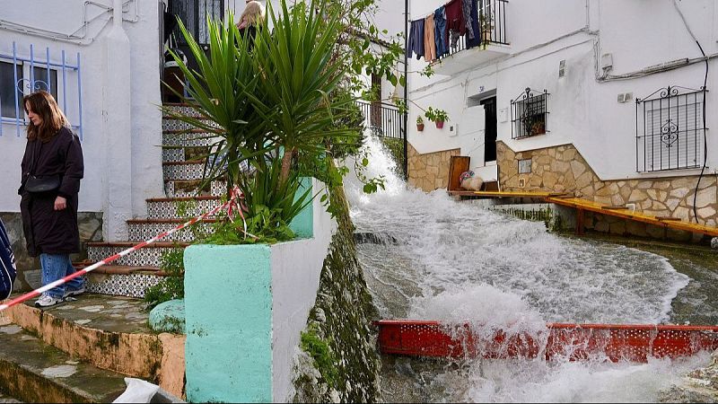 Calle empinada de pueblo inundada por un torrente de agua, con una pasarela de madera y una mujer observando desde una escalinata.