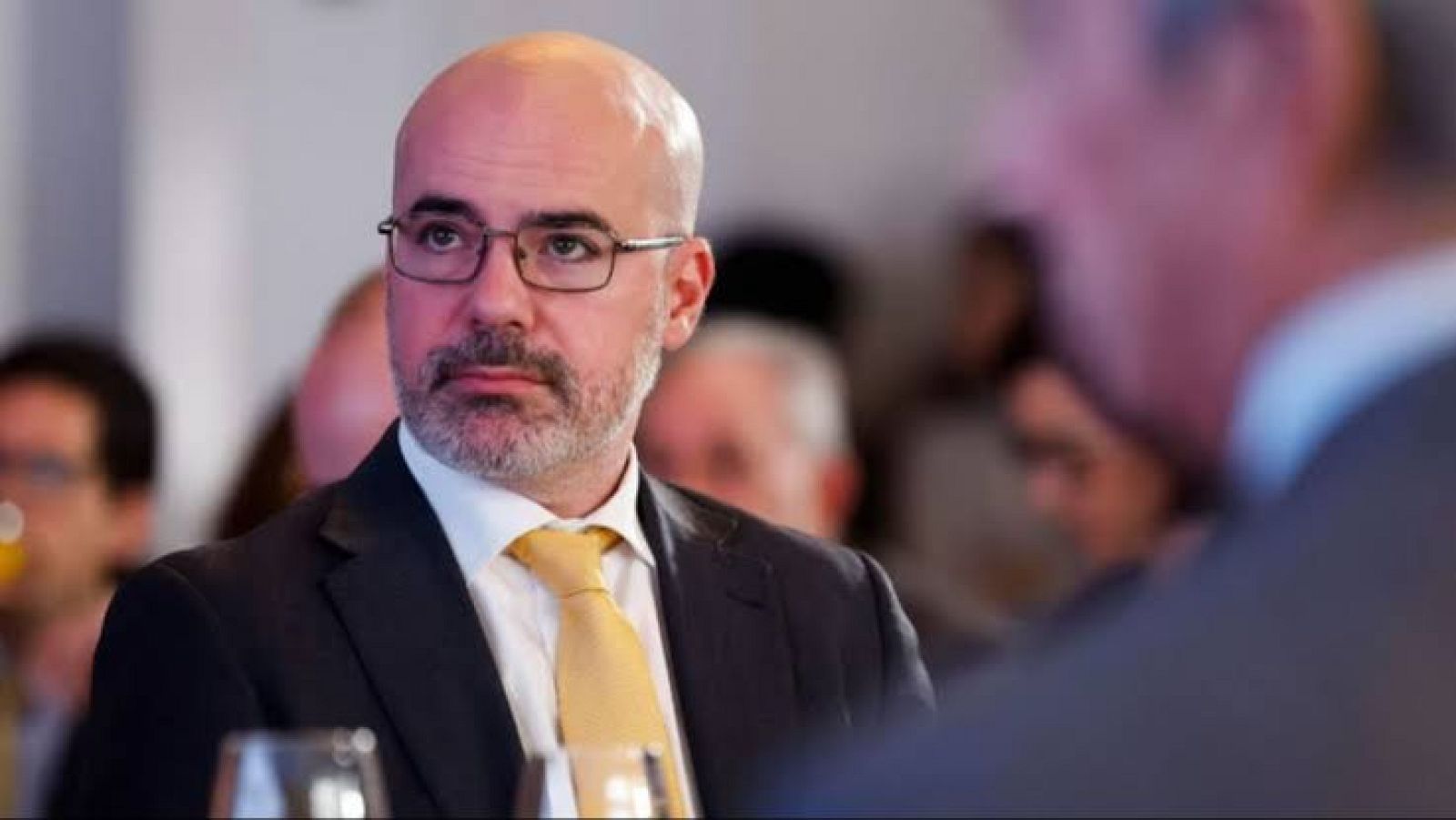 Plano medio corto del político Jorge Buxadé con chaqueta oscura, camisa blanca y corbata amarilla, mostrando expresión seria en un evento institucional.