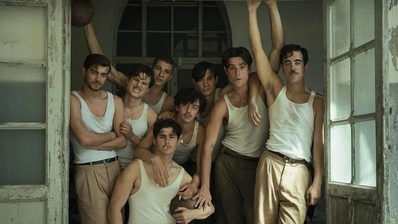 Grupo de ocho jóvenes con indumentaria de época, camisetas de tirantes y pantalones de talle alto, posando con balones de cuero clásico en una puerta antigua.