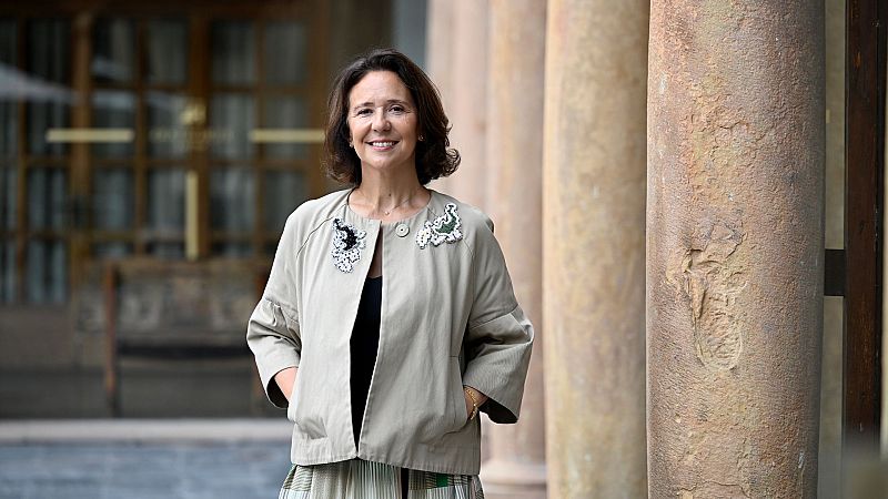 Teresa Sanjurjo posa sonriente en el patio de un edificio histórico, vistiendo una chaqueta beige con apliques bordados y columnas de piedra de fondo.