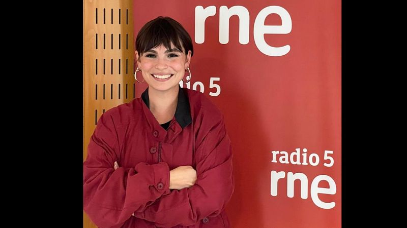 La escritora Andrea Abreu posa sonriente en el estudio de radio con chaqueta burdeos y el logotipo de la emisora al fondo.