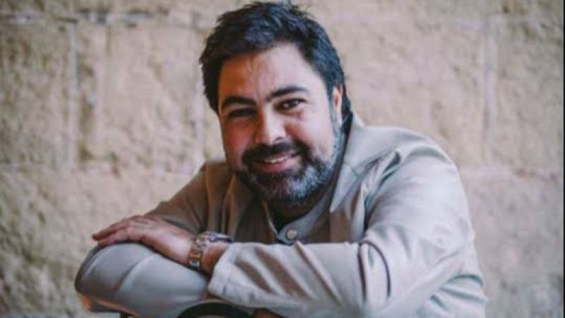 Plano medio del cantaor Antonio Campos sonriendo relajado, apoyado en una silla frente a una pared de piedra.
