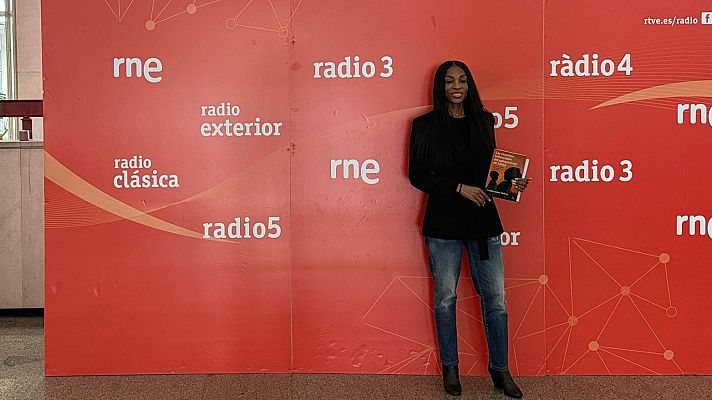 Guillermina Mekuy, con chaqueta negra y jeans, posa frente a una pared naranja con logotipos de emisoras de radio, sosteniendo un libro.