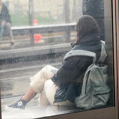 Una persona con chaqueta oscura y mochila espera sentada en una marquesina de cristal mojada por la lluvia.