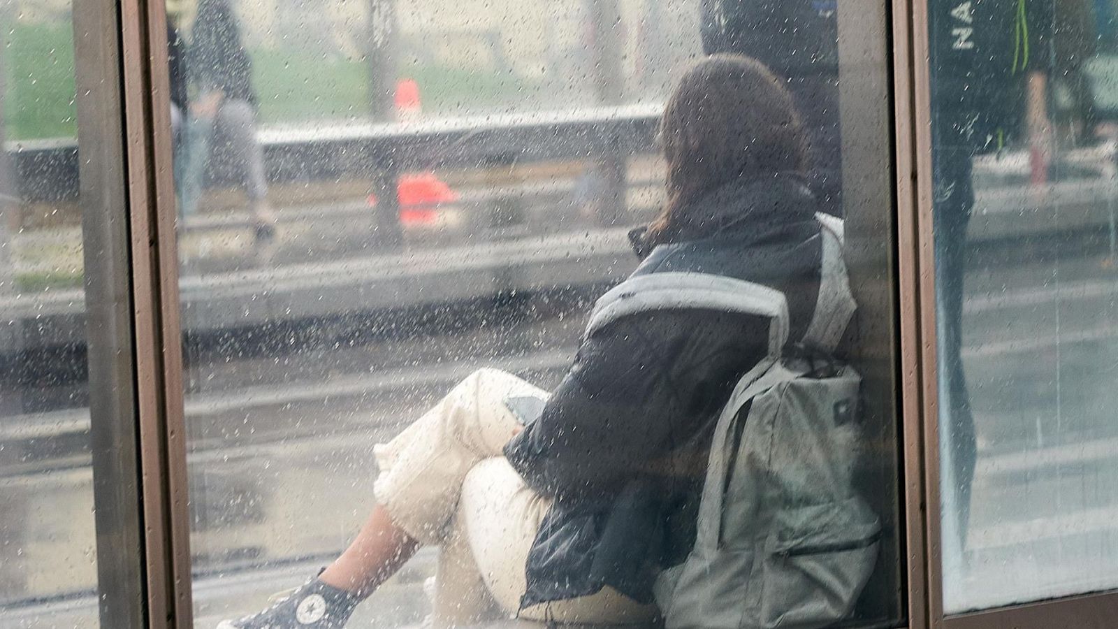Una persona con chaqueta oscura y mochila espera sentada en una marquesina de cristal mojada por la lluvia.
