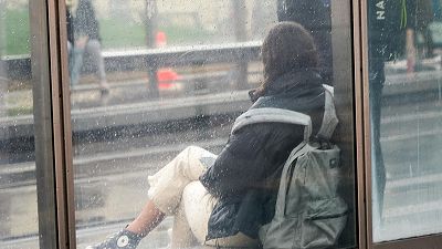 Una persona con chaqueta oscura y mochila espera sentada en una marquesina de cristal mojada por la lluvia.