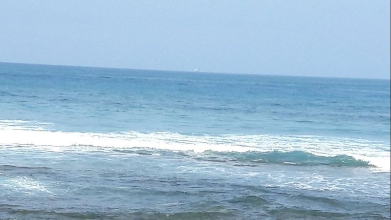 Paisaje marino con cielo despejado, mar en tonos azules y turquesas, una ola rompiendo con espuma blanca y un fondo rocoso en primer plano.