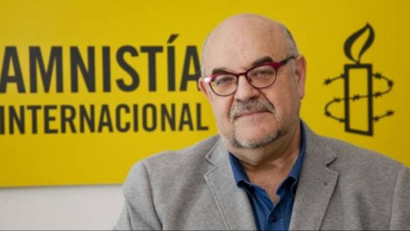 Esteban Beltrán, director de Amnistía Internacional en España, con gafas y barba corta, posa sonriente ante un fondo amarillo con el logotipo de la organización.