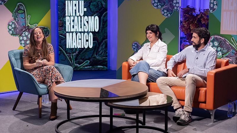 Plat� de entrevistas con una presentadora en sill�n azul y dos invitados en sof� naranja frente a una mesa redonda, con fondo luminoso y decoraci�n colorida.