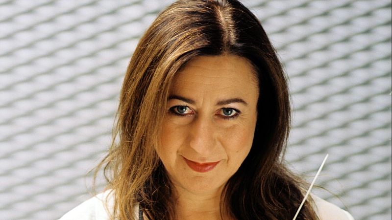 Retrato en primer plano de la directora Inma Shara sonriendo levemente, sosteniendo una batuta sobre un fondo gris desenfocado.