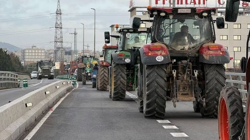 Caravana de tractores agr�colas circulando en marcha lenta por una carretera junto a una barrera de hormig�n, con un paisaje urbano y cielo nublado al fondo.
