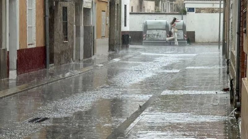 Calle estrecha y adoquinada completamente inundada por una lluvia torrencial, con edificios antiguos a los lados y una persona cruzando corriendo al fondo.
