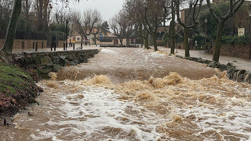 Caudal torrencial de agua turbia desbordando una riera urbana con muros de piedra, aceras y �rboles bajo un cielo nublado.