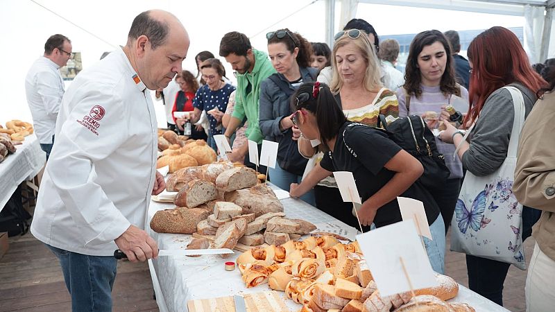 Un panadero corta hogazas de pan artesanal en una mesa llena de productos y carteles, ante la atenta mirada del público en una feria gastronómica.