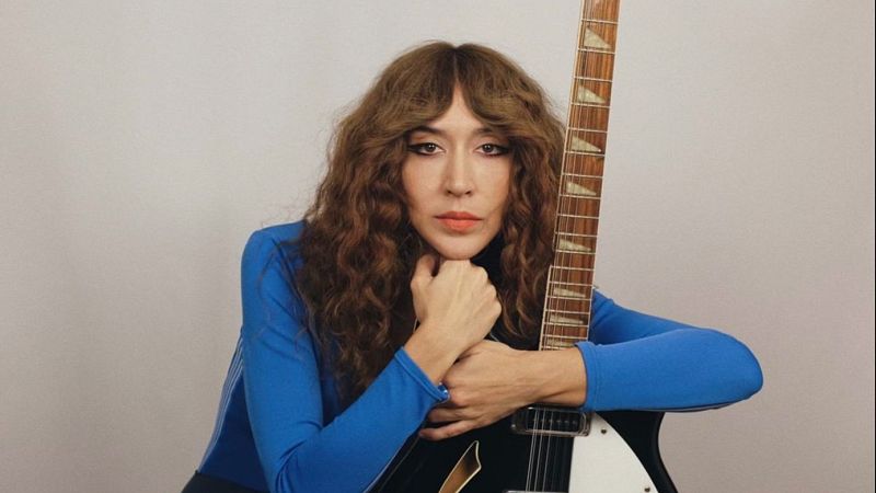 Mujer de pelo castaño con flequillo y camiseta azul eléctrico posa en plano medio sosteniendo una guitarra eléctrica sobre fondo gris.