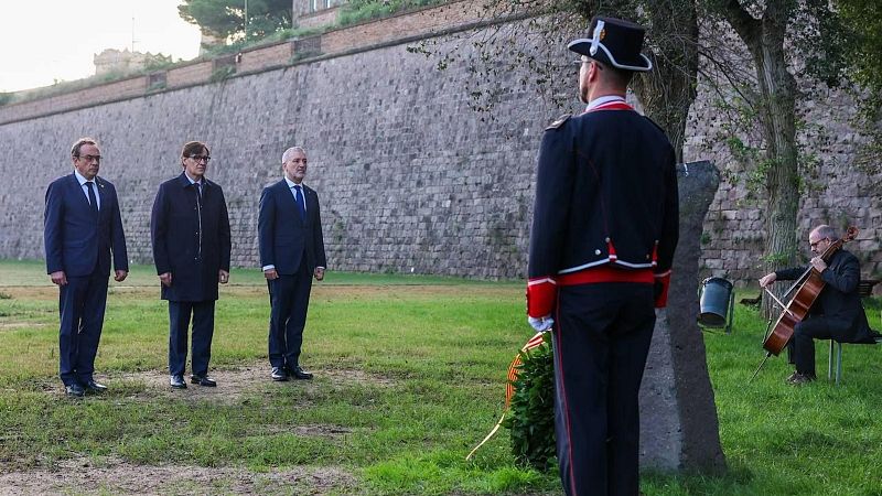 Autoridades políticas participan en un homenaje con ofrenda floral junto a un monolito y un agente de los Mossos d'Esquadra en una fortaleza histórica.