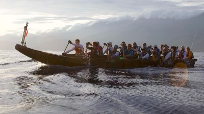 Expedición de remeros en una gran canoa tradicional de madera navegando por aguas abiertas frente a una costa montañosa con cordillera nublada al fondo.