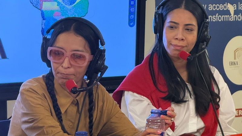 Dos mujeres con auriculares y micrófonos en un estudio de radio; una lleva trenzas y gafas rosadas, y la otra sostiene una botella de agua.