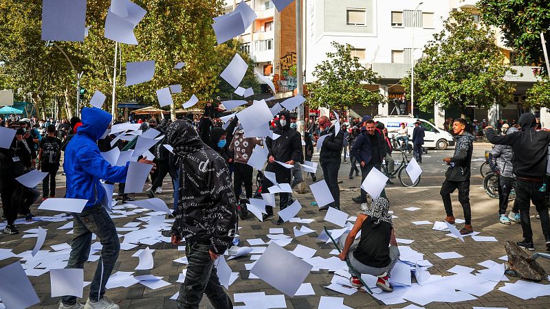 Protesta urbana en una calle adoquinada con manifestantes lanzando hojas de papel al aire rodeados de edificios y árboles.