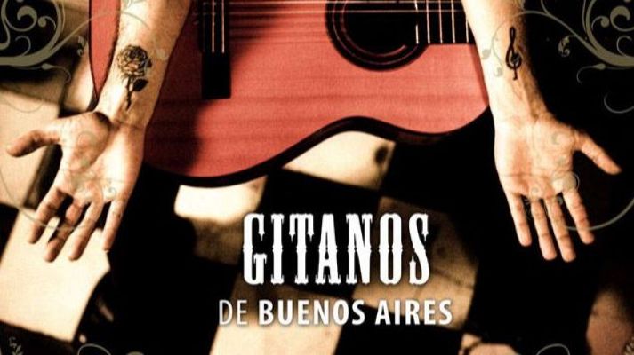 Vista cenital de un músico sentado con los brazos abiertos sobre suelo ajedrezado, mostrando tatuajes en las muñecas y una guitarra roja arriba.