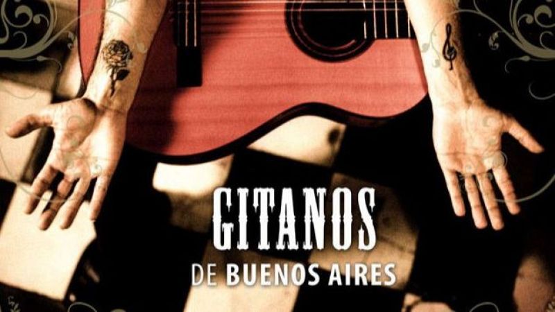 Vista cenital de un músico sentado con los brazos abiertos sobre suelo ajedrezado, mostrando tatuajes en las muñecas y una guitarra roja arriba.