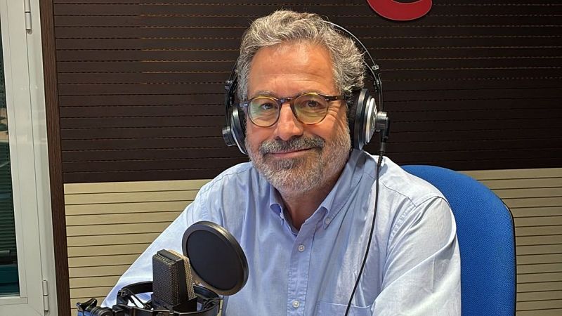 El periodista Josep Cuní sonríe en un estudio de radio con auriculares y micrófono profesional, luciendo camisa azul y gafas.