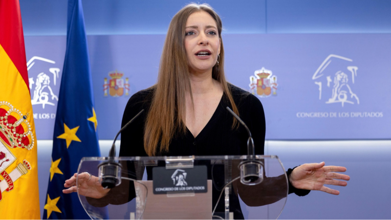 Plano medio de la pol�tica Ester Mu�oz tras un atril transparente en una comparecencia, con expresi�n seria y gesto explicativo ante las banderas oficiales.