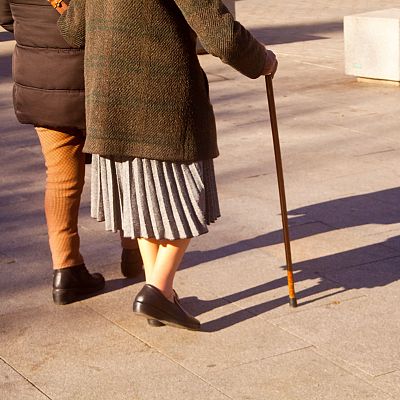 Dos personas caminan por una plaza adoquinada bajo una luz de atardecer, una de ellas apoyándose en un bastón y vistiendo abrigo oscuro y falda plisada.