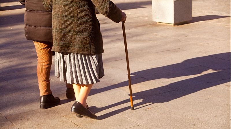 Dos personas caminan por una plaza adoquinada bajo una luz de atardecer, una de ellas apoyándose en un bastón y vistiendo abrigo oscuro y falda plisada.