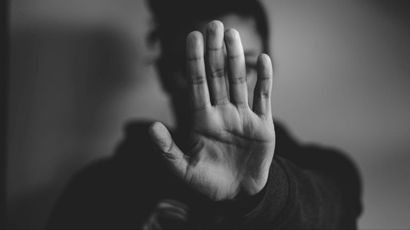 Fotograf�a en blanco y negro de una mano abierta en primer plano haciendo el gesto de alto, con una persona con capucha desenfocada al fondo.