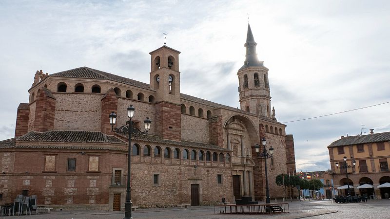 Vista exterior diurna de una iglesia monumental con torre y chapitel junto a una plaza adoquinada con farolas cl�sicas y edificios con soportales.
