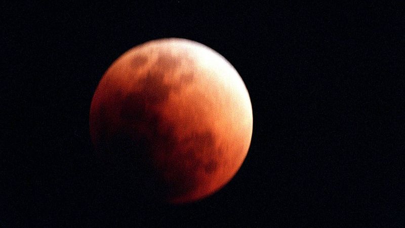 Eclipse lunar con la Luna en tonos rojo cobrizo y una franja curva muy brillante en el borde superior derecho sobre un fondo negro.