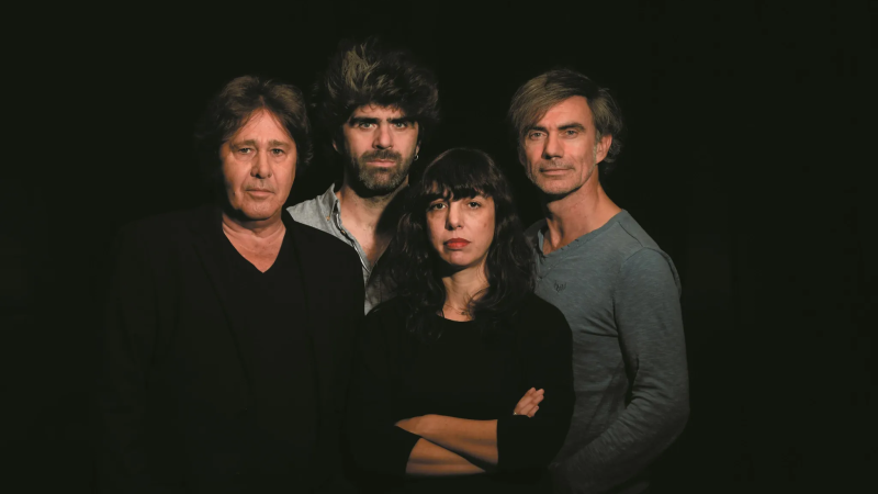 Retrato en plano medio de un cuarteto musical de tres hombres y una mujer con ropa oscura sobre fondo negro y luz dram�tica.