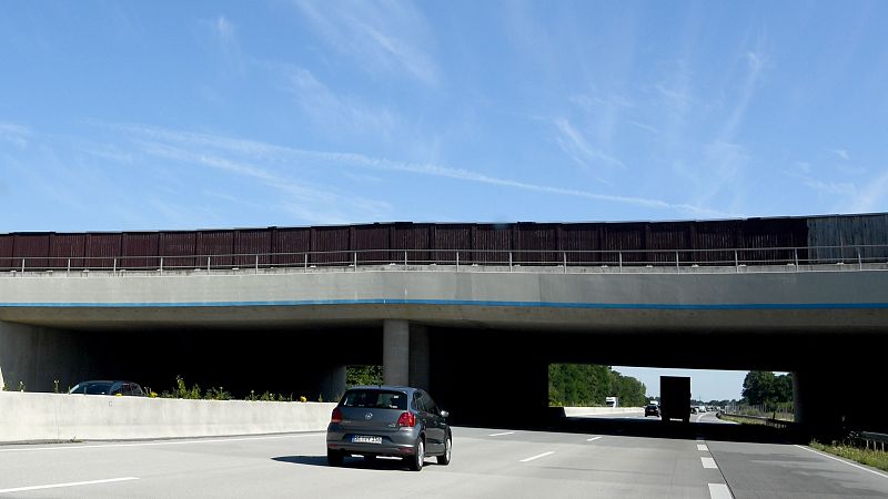 Coche gris circulando por una autopista bajo un puente elevado para animales con paneles protectores oscuros.