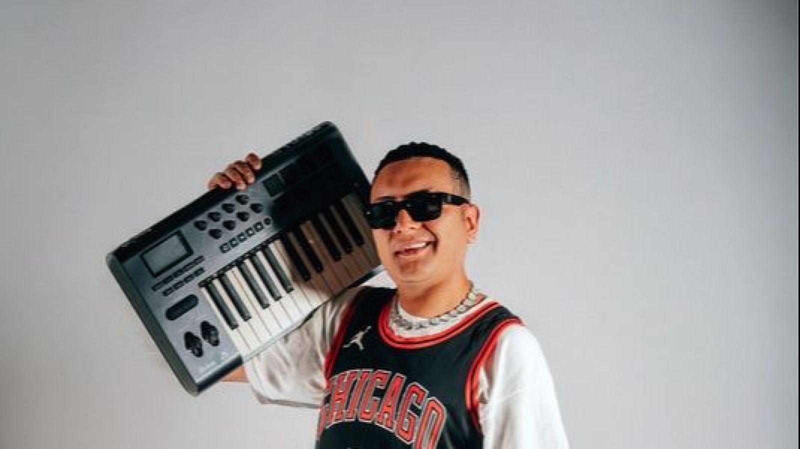 El artista musical DJ Sue�o posa en plano medio con estilo urbano, gafas de sol y un teclado MIDI apoyado en el hombro.